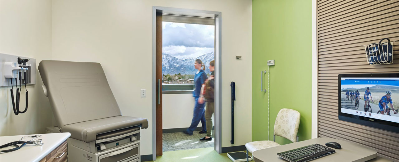 Modern examination room with an open sliding door, green wall, medical equipment, and a monitor displaying an image