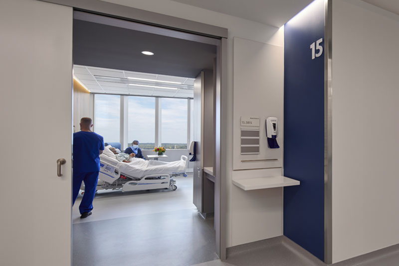 Open sliding door with a nurse and patient inside a bright hospital room with a hand sanitizer station