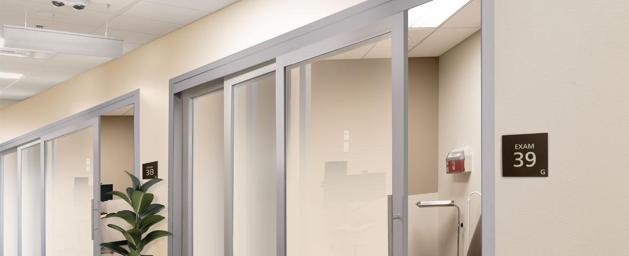 Rendering of a healthcare corridor with frosted glass telescopic sliding doors leading to examination rooms, enhancing privacy and accessibility.