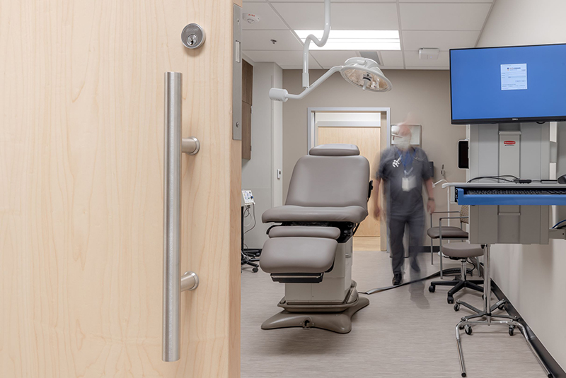Close-up of a wooden door with stainless steel hardware leading into a medical exam room with an examination chair and computer station.