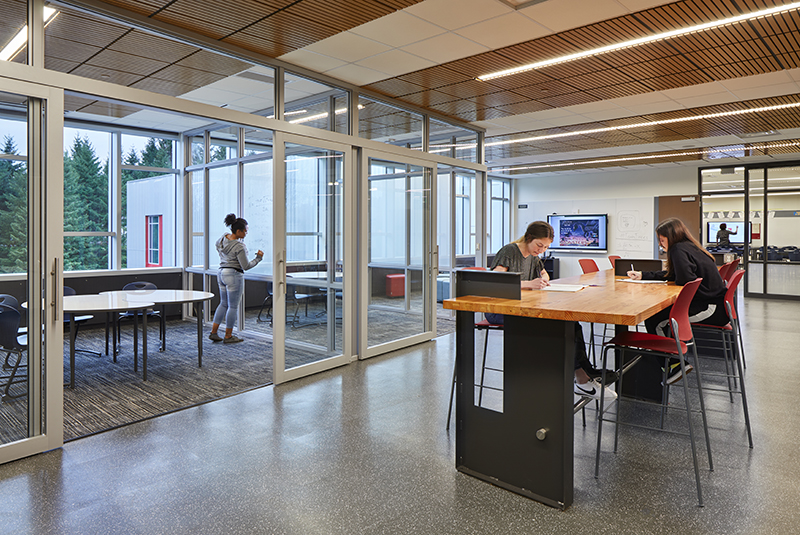 School space with women collaborating at table in the center with sliding glass doors leading to private rooms on the exterior walls.